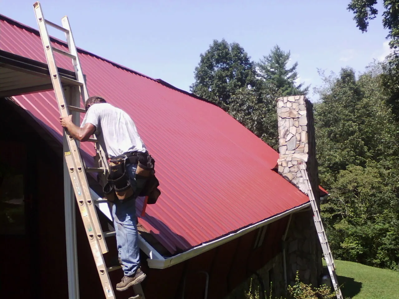 Roofer on a ladder beside a red metal roof and stone chimney — Soffit & Fascia Repair by Roofing Brothers in Luckey